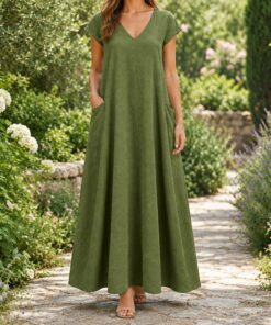 Woman in an olive linen dress with hands in deep pockets among garden plants, shown from the neck down
